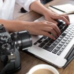 Hands typing on laptop next to camera – digital creator planning Onlyfans job application or side gig work